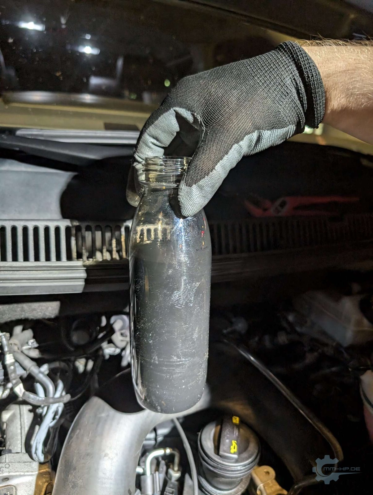 Gloved hand holding a bottle of dark oil condensate above the engine bay