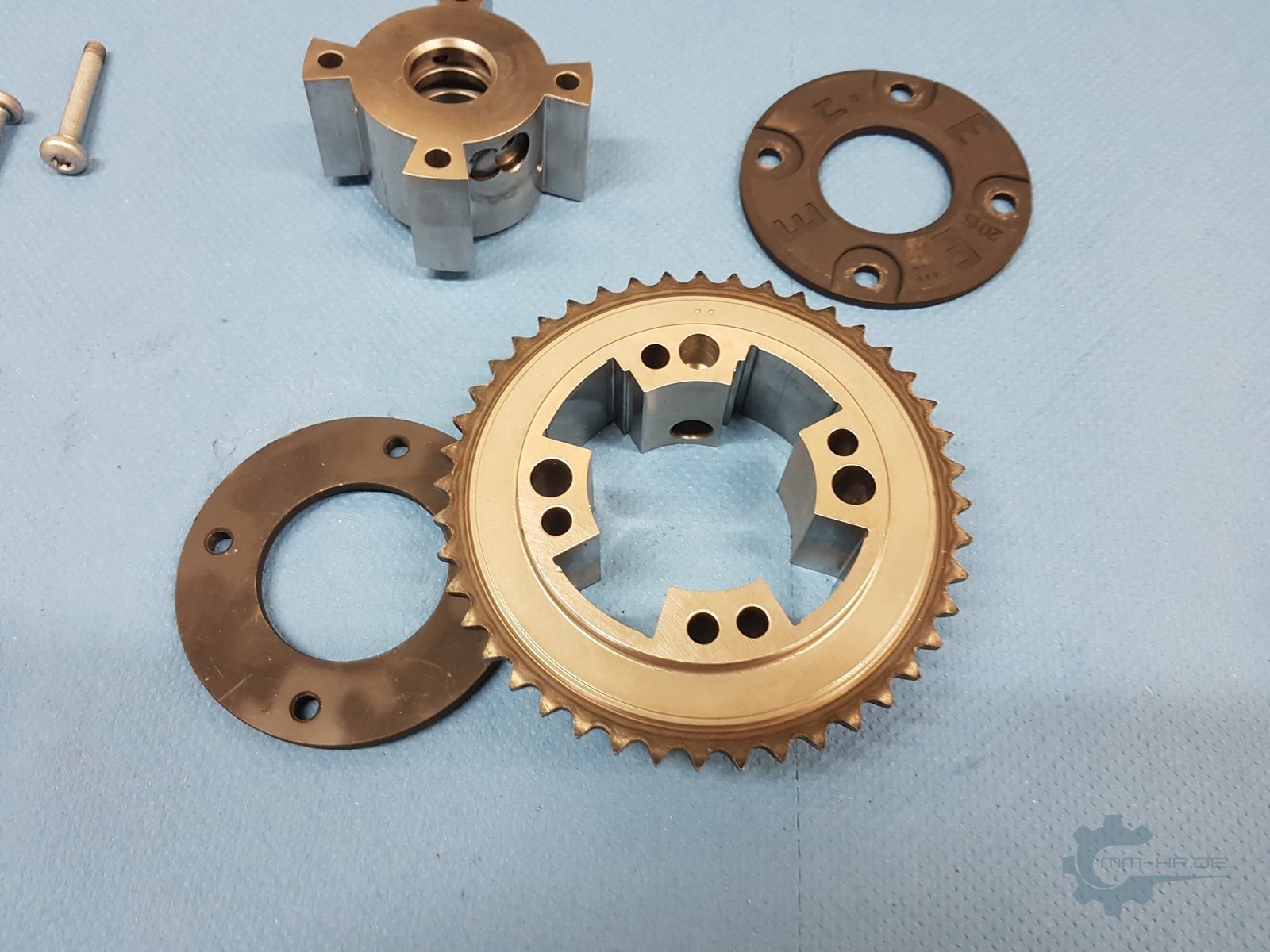 A disassembled cam phaser on blue workshop paper: housing with chain sprocket and chambers, rotor with vanes, two cover discs and screws